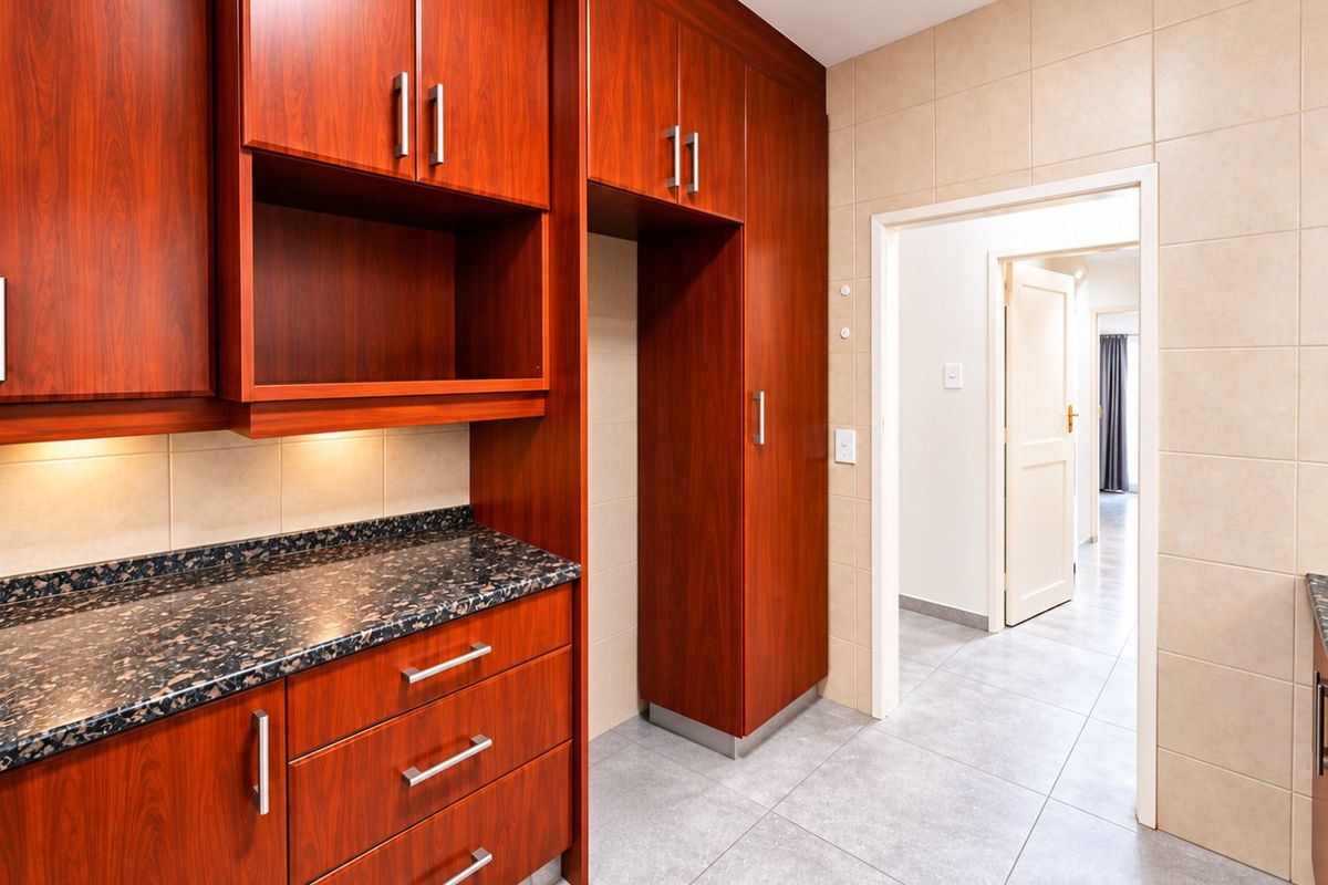 A clean kitchen area featuring warm-toned wood cabinetry, dark granite countertops, and neutral tiled flooring, leading into a hallway.