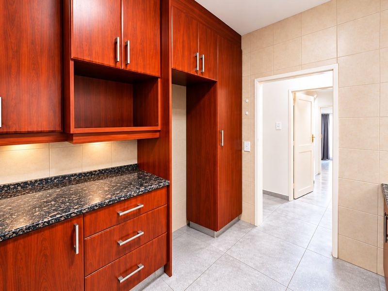 A clean kitchen area featuring warm-toned wood cabinetry, dark granite countertops, and neutral tiled flooring, leading into a hallway.