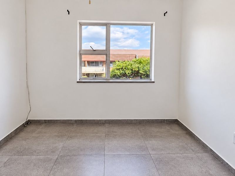 A bright, empty bedroom featuring neutral tiled flooring and a large window, providing a clean and versatile space ready for personalization.