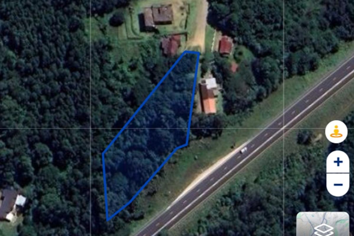 An aerial satellite view showing a residential land plot outlined in blue, situated near a main road and surrounded by dense vegetation.