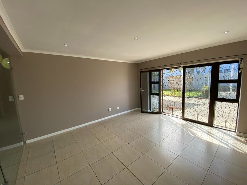 A bright, empty living room featuring neutral tiled flooring, recessed downlighting, and large sliding glass doors with decorative security gates leading to the garden.