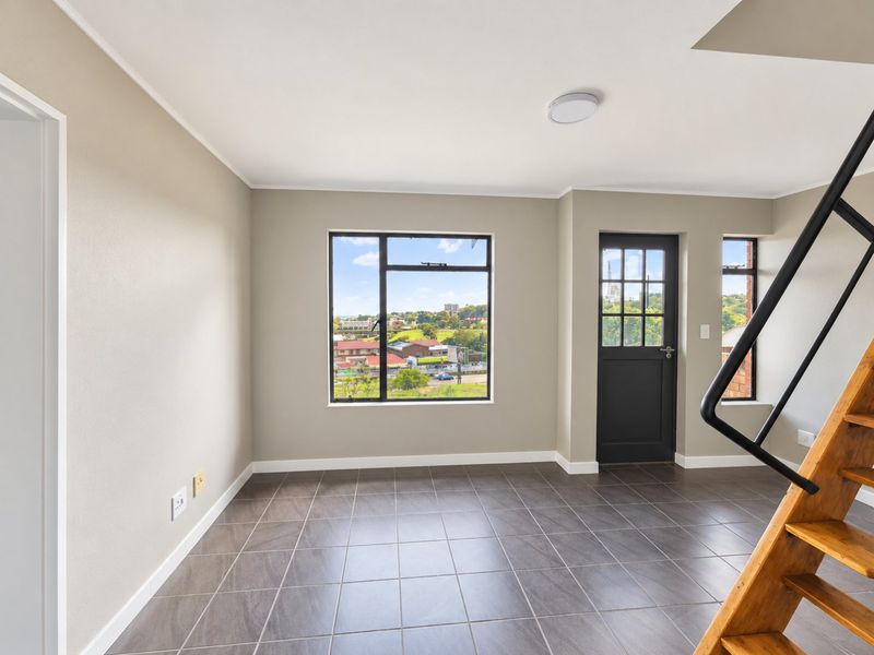 A bright, empty living area featuring modern tiled flooring, a wooden staircase with a black handrail, and large windows offering an elevated neighborhood view.