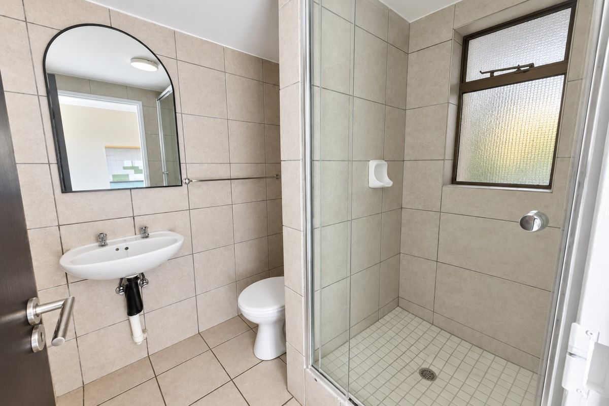 A functional bathroom featuring a glass-enclosed shower, wall-mounted basin, and toilet, finished with neutral floor-to-ceiling tiling and an obscured glass window.