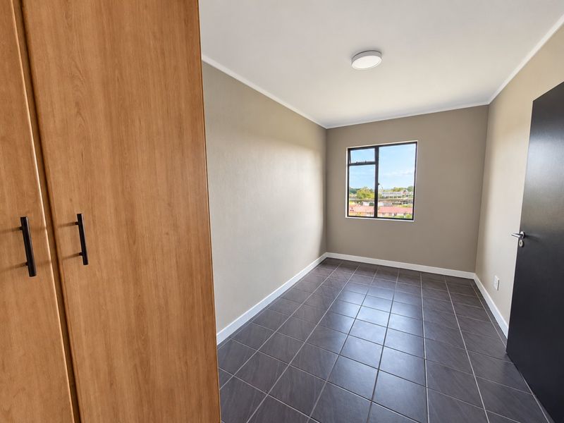 A clean, empty bedroom featuring neutral wall tones, dark tiled flooring, and a built-in wooden wardrobe, offering a bright and versatile living space.