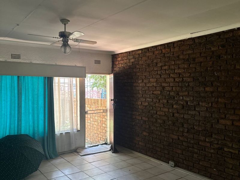 Spacious living area featuring a rustic exposed brick wall, tiled flooring, and a ceiling fan, with direct access to an outdoor area via a security gate.