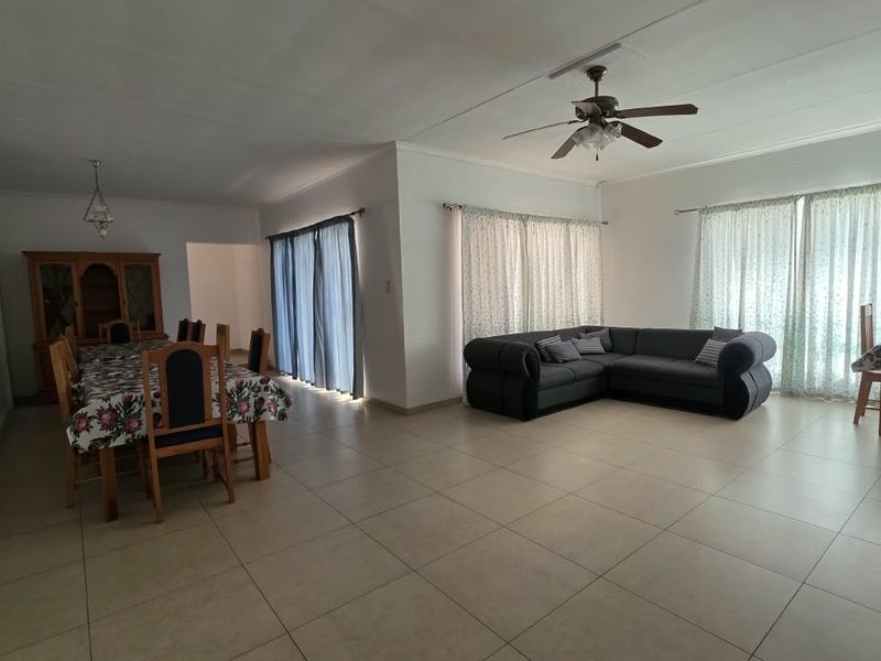 Spacious open-plan living and dining area featuring neutral tiled flooring, large windows with curtain rails, and a central ceiling fan.