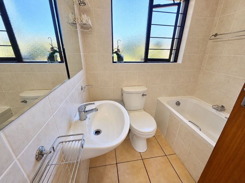 A clean and functional bathroom featuring neutral ceramic wall and floor tiles, a built-in bathtub, pedestal washbasin, and a large wall mirror.