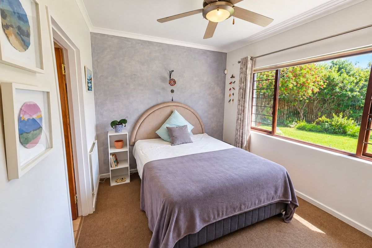 A well-lit bedroom featuring a textured accent wall, ceiling fan, and large window overlooking the garden.