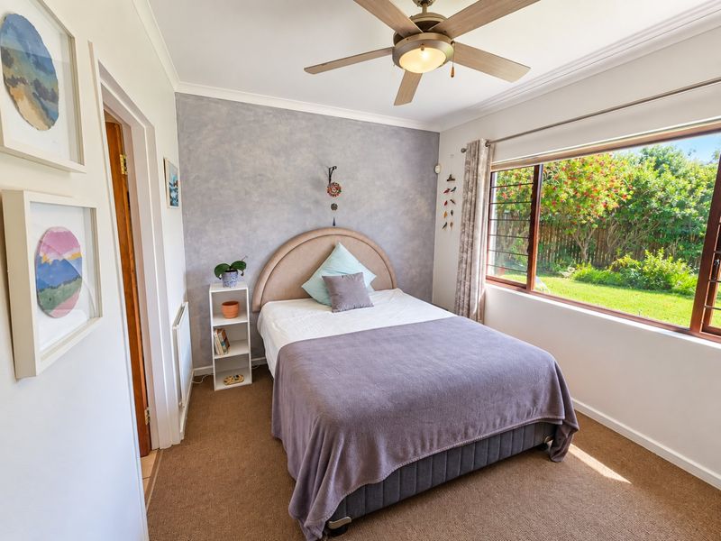 A well-lit bedroom featuring a textured accent wall, ceiling fan, and large window overlooking the garden.