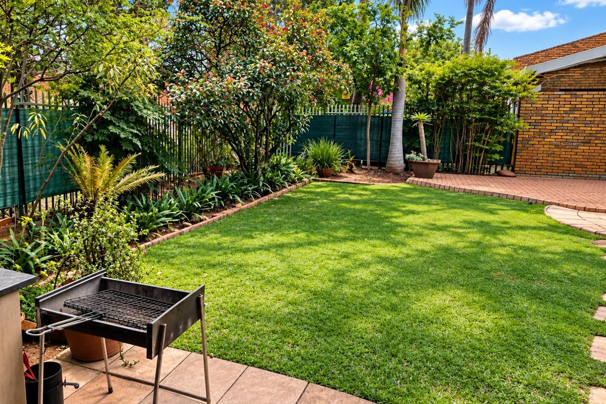 A well-maintained private rear garden featuring a paved patio area, lush green lawn, and established perimeter landscaping with brick and metal fencing.