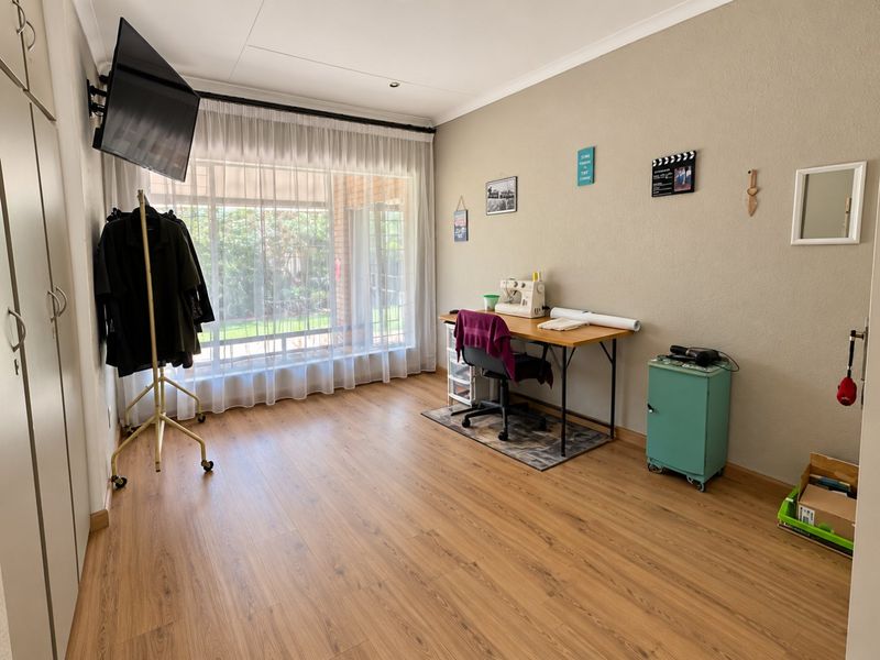 A bright bedroom featuring laminate flooring, built-in cupboards, and a large window with sheer curtains, offering a versatile space for a bedroom or office.