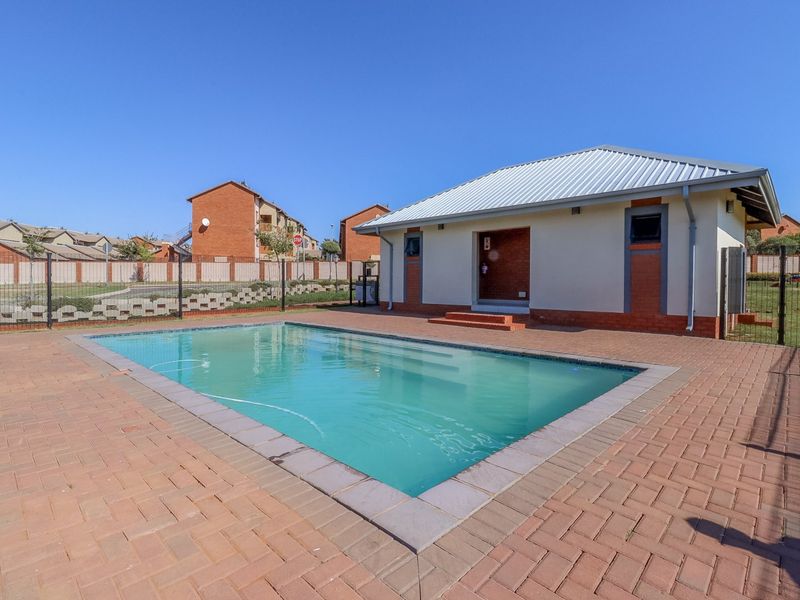 A well-maintained communal swimming pool area featuring paved decking and a brick-built clubhouse structure within a secure residential complex.