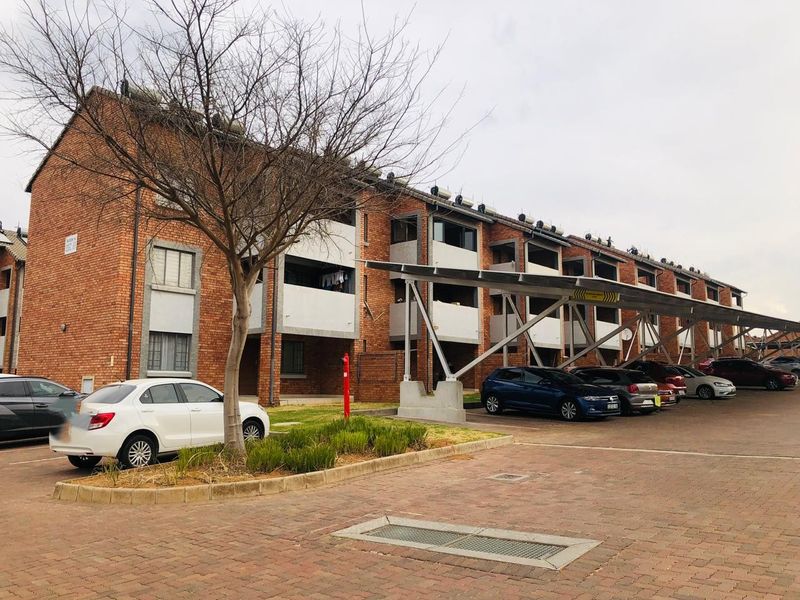 Exterior view of a multi-story brick apartment complex featuring covered parking bays, paved driveway, and solar water heating units on the roof.