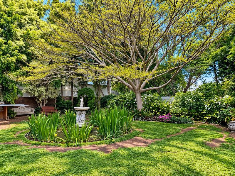 A lush, well-maintained garden featuring a central stone fountain, mature trees, and a brick pathway set within a landscaped green space.