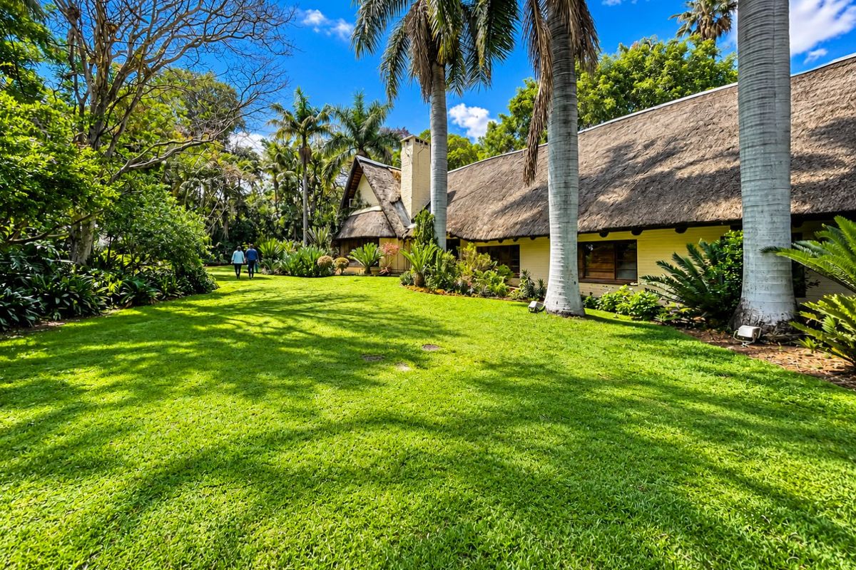 A spacious rear garden featuring a well-maintained lawn, mature palm trees, and a traditional house with a classic thatch roof.