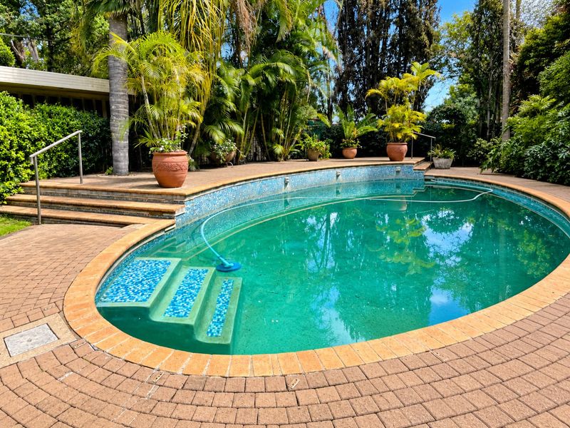 A well-maintained outdoor swimming pool area featuring brick paving, integrated pool steps, and a metal handrail, surrounded by lush garden landscaping.