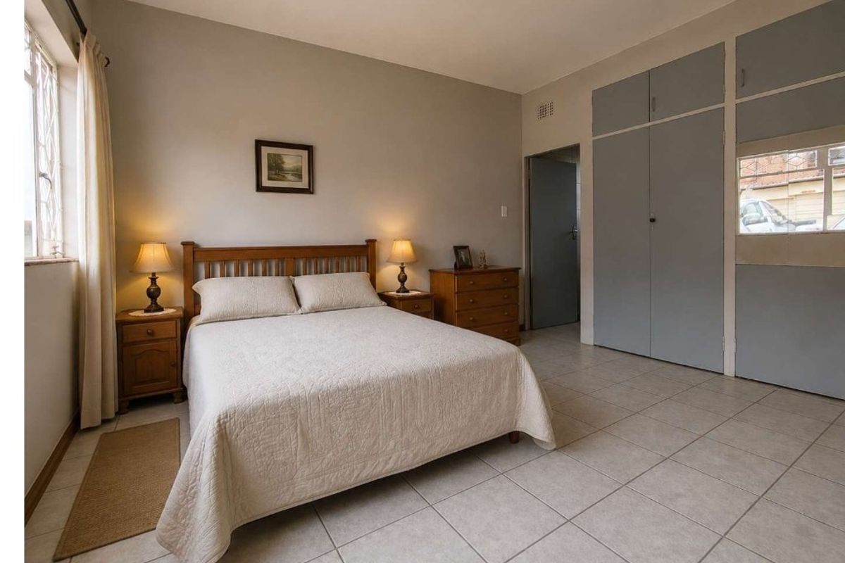 A well-lit bedroom featuring neutral tiled flooring, built-in cupboards, and a window with security bars, offering a functional and comfortable living space.