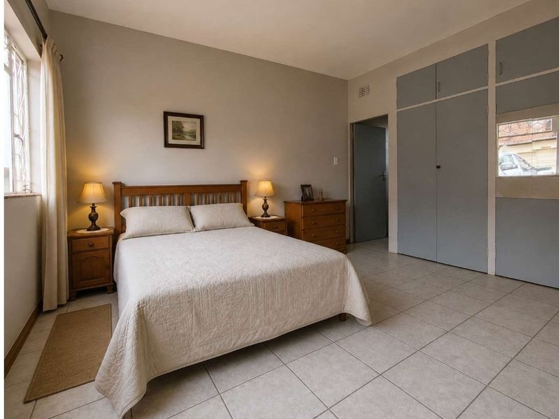A well-lit bedroom featuring neutral tiled flooring, built-in cupboards, and a window with security bars, offering a functional and comfortable living space.