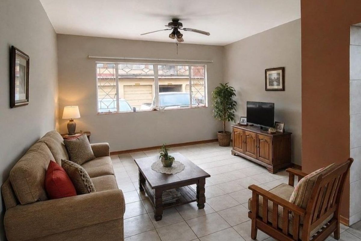 A comfortable living room featuring tiled flooring, a ceiling fan, and a large window with security bars, providing natural light.