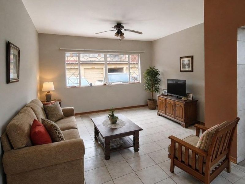 A comfortable living room featuring tiled flooring, a ceiling fan, and a large window with security bars, providing natural light.