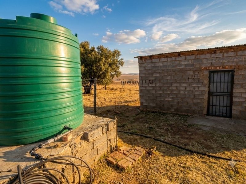 Exterior view of a farm outbuilding featuring a large green water storage tank on a concrete base and a simple brick structure.