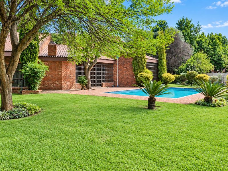 A well-maintained backyard featuring a swimming pool, brick-paved patio area, and established garden landscaping set against a classic brick house exterior.