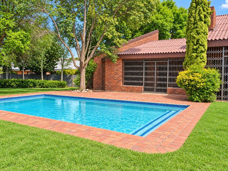 A well-maintained swimming pool area featuring brick paving, established garden surroundings, and a brick-built home exterior with security gates.
