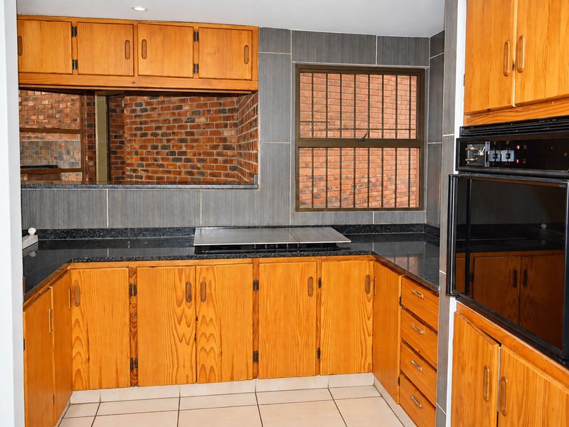 A functional kitchen featuring wooden cabinetry, granite countertops, built-in oven and hob, and a unique exposed brick feature wall with security-barred window.