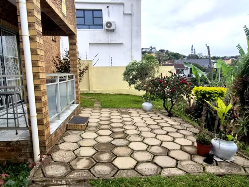 Rear exterior view featuring a paved patio area, brick facade, and sliding glass doors with security gates, overlooking a landscaped garden.