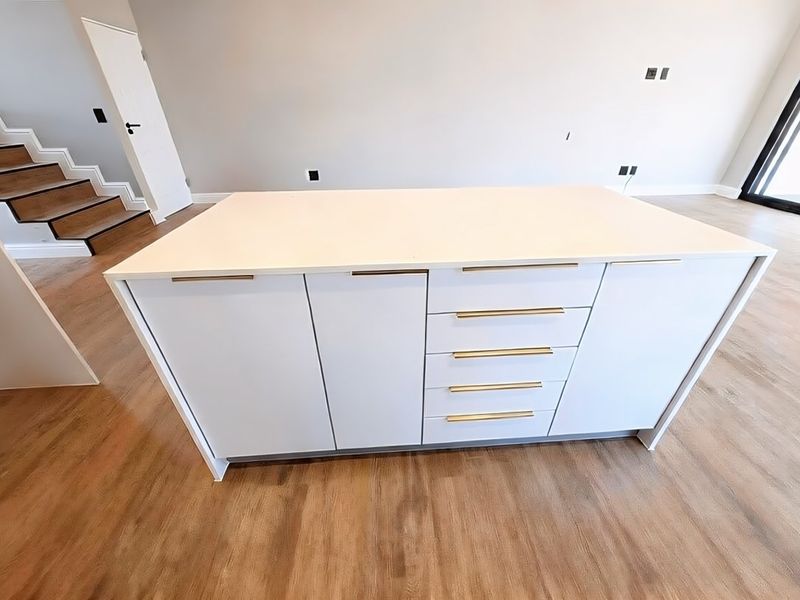 A modern kitchen island with white cabinetry and gold hardware, set in an open-plan space with wood-look flooring and an adjacent staircase.