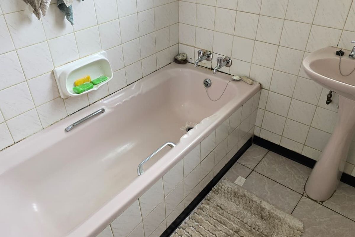 A dated bathroom featuring a pink bathtub and matching pedestal washbasin, complemented by white wall tiles and neutral floor tiling.