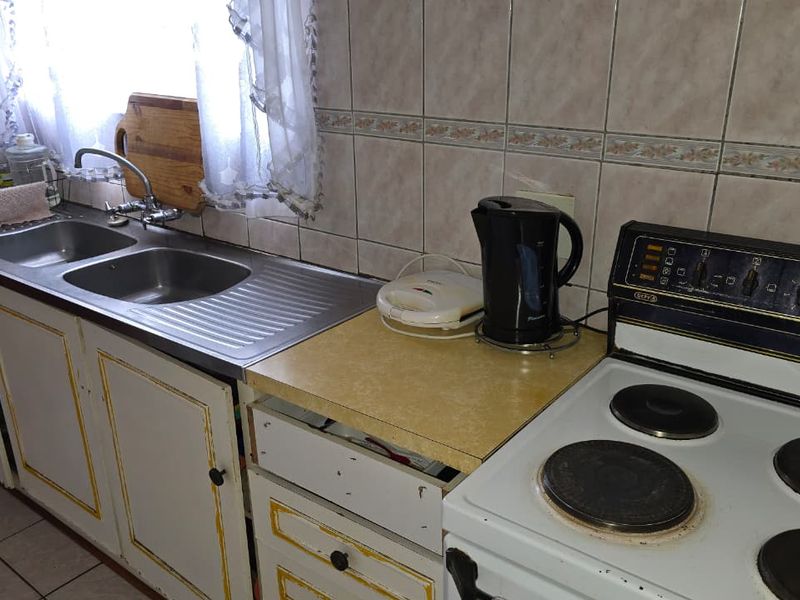 A functional kitchen featuring a double bowl stainless steel sink, tiled splashback, and laminate countertops, currently showing signs of wear and missing cabinetry components.
