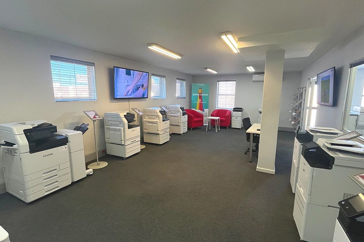 A spacious commercial office area featuring carpeted flooring, multiple windows with blinds, and integrated lighting, currently utilized as a showroom for office equipment.