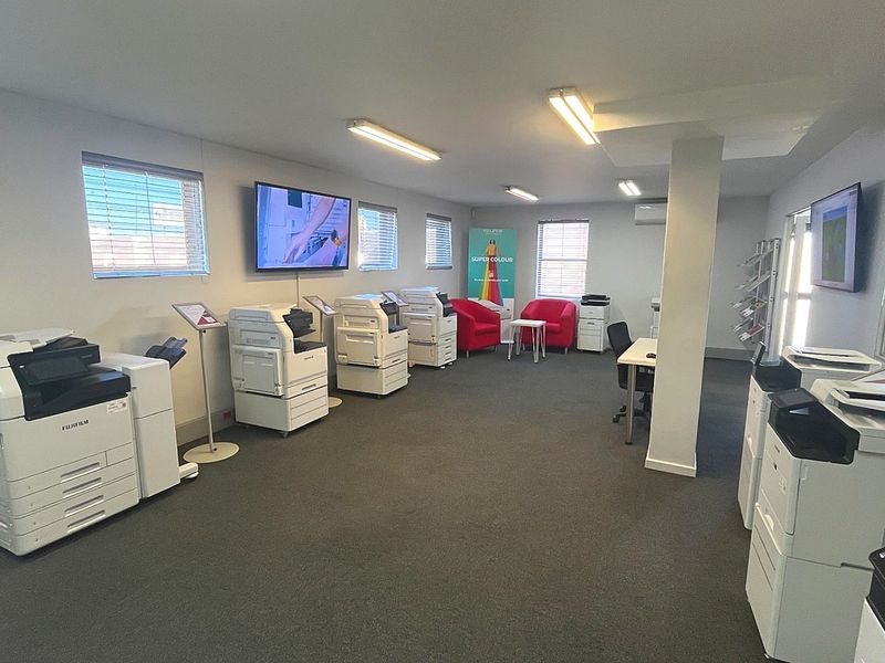 A spacious commercial office area featuring carpeted flooring, multiple windows with blinds, and integrated lighting, currently utilized as a showroom for office equipment.