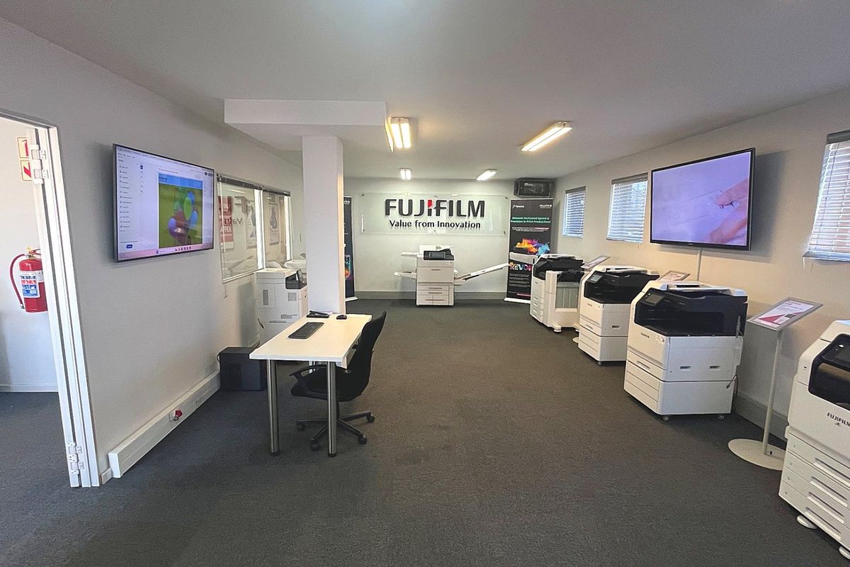 A well-maintained commercial office space featuring carpeted flooring, wall-mounted digital displays, and professional lighting, currently utilized as a showroom for office equipment.