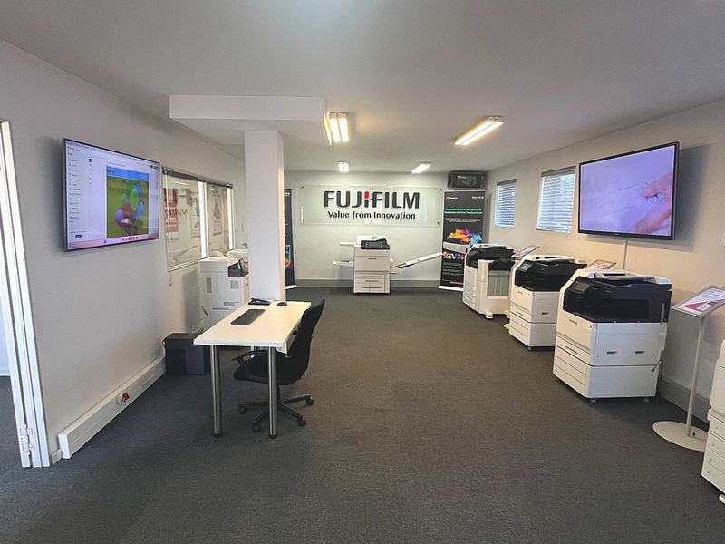 A well-maintained commercial office space featuring carpeted flooring, wall-mounted digital displays, and professional lighting, currently utilized as a showroom for office equipment.