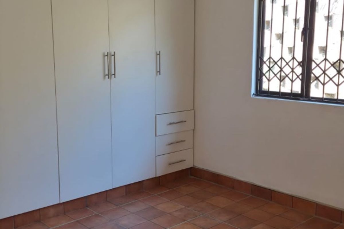 A clean, empty bedroom featuring built-in cupboards, terracotta-style ceramic floor tiles, and a window fitted with a security gate.