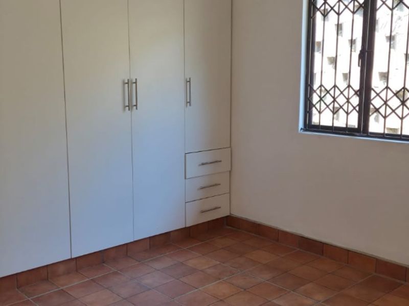 A clean, empty bedroom featuring built-in cupboards, terracotta-style ceramic floor tiles, and a window fitted with a security gate.