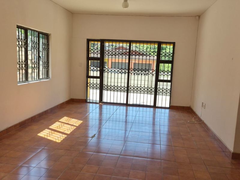 A spacious, empty living room featuring tiled flooring, security gates on the windows, and a sliding glass door leading to the exterior.
