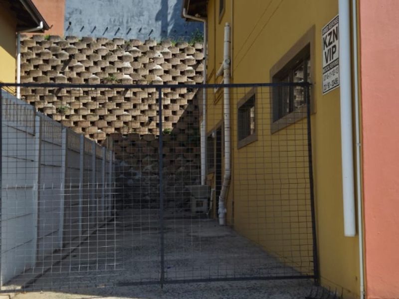 A narrow rear exterior area featuring a concrete paved surface, a retaining wall, and a metal security gate.
