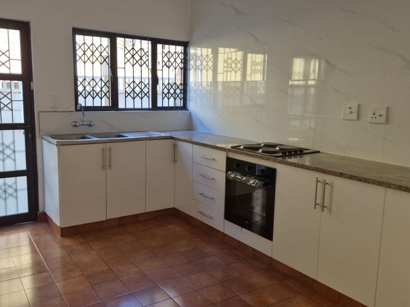 A clean, functional kitchen featuring white cabinetry, granite countertops, a built-in oven and hob, and tiled splashbacks with secure window and door access.