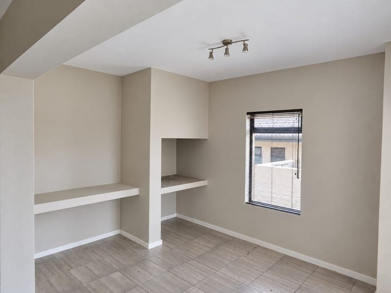 A bright, empty living area featuring neutral tiled flooring, built-in shelving, a recessed alcove, and a window with fitted blinds.