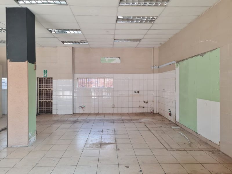 An empty, open-plan commercial space featuring tiled flooring, a suspended ceiling with recessed lighting, and a tiled wall section, requiring cosmetic refurbishment.
