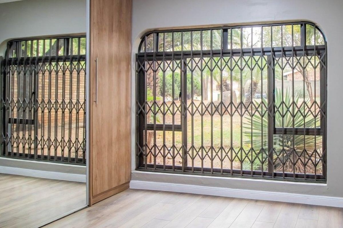 A bright bedroom featuring modern laminate flooring, a large built-in wardrobe with mirrored sliding doors, and secure metal security gates on the windows.