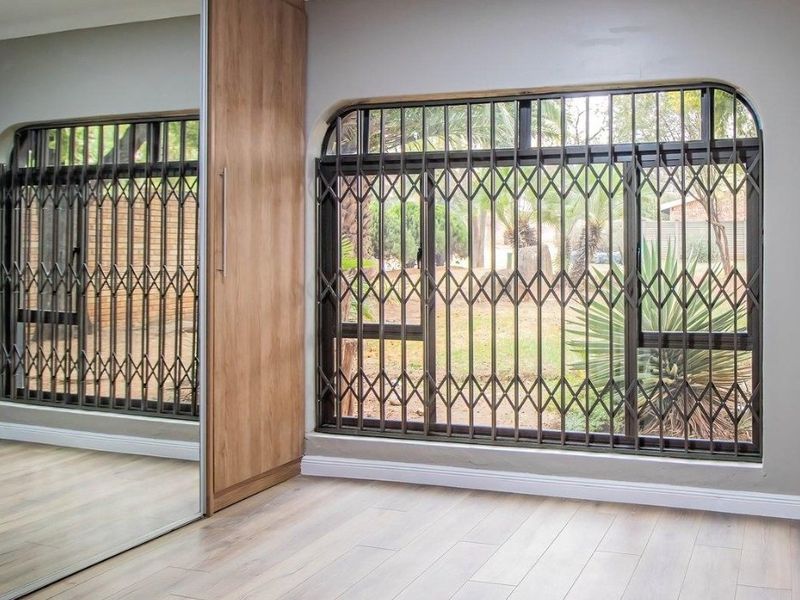 A bright bedroom featuring modern laminate flooring, a large built-in wardrobe with mirrored sliding doors, and secure metal security gates on the windows.
