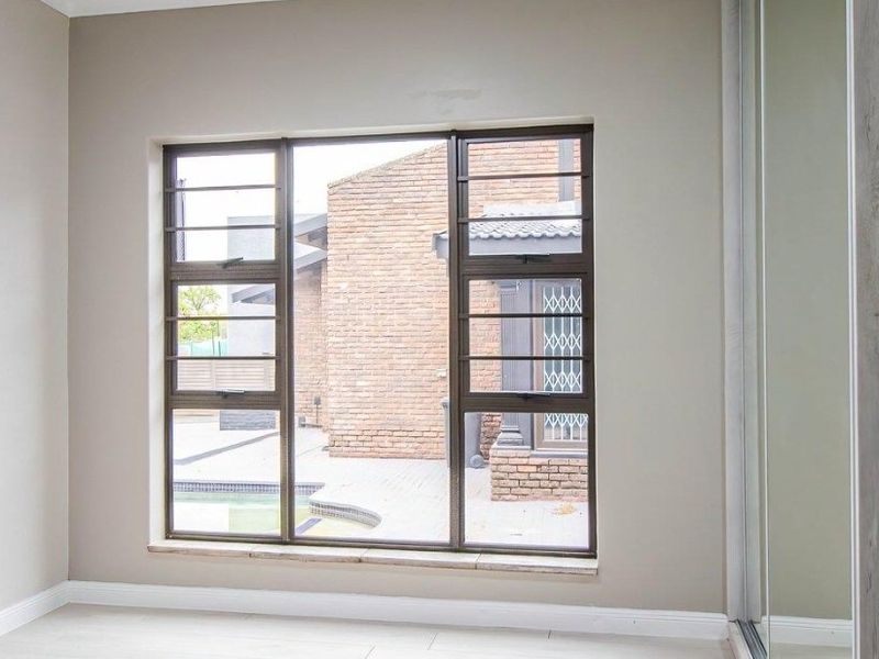 A bright, empty bedroom featuring neutral walls, light flooring, a built-in sliding wardrobe, and large windows with security bars overlooking the exterior area.