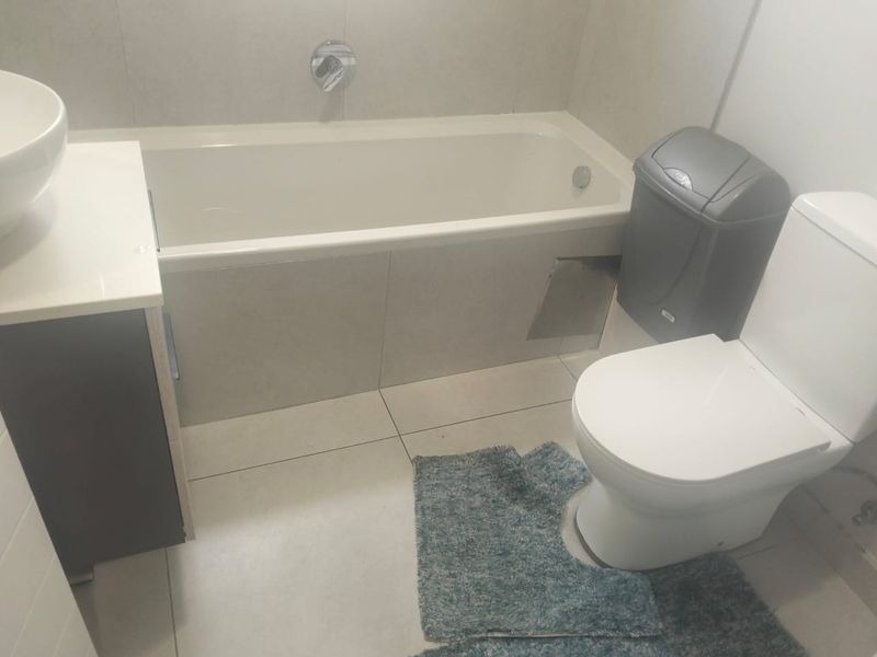 A compact bathroom featuring a bathtub, toilet, and vanity basin with neutral wall and floor tiling. Requires repair to the bathtub apron.