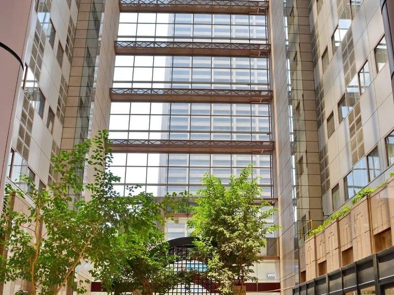 A grand, light-filled atrium entrance featuring a soaring glass ceiling, stone flooring, and integrated indoor planters within a modern apartment complex.