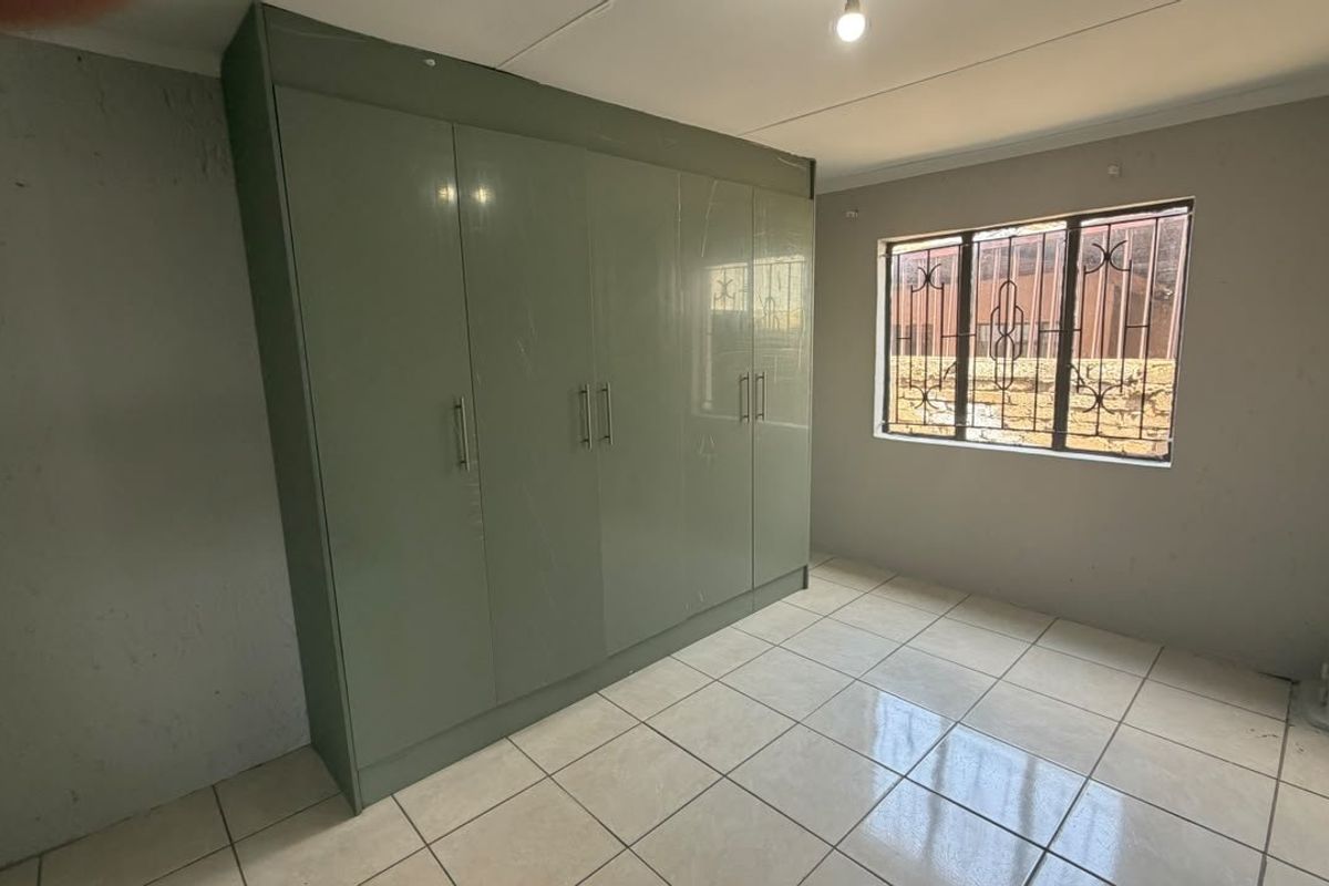 A vacant bedroom featuring tiled flooring, built-in cupboards, and a window fitted with security bars.