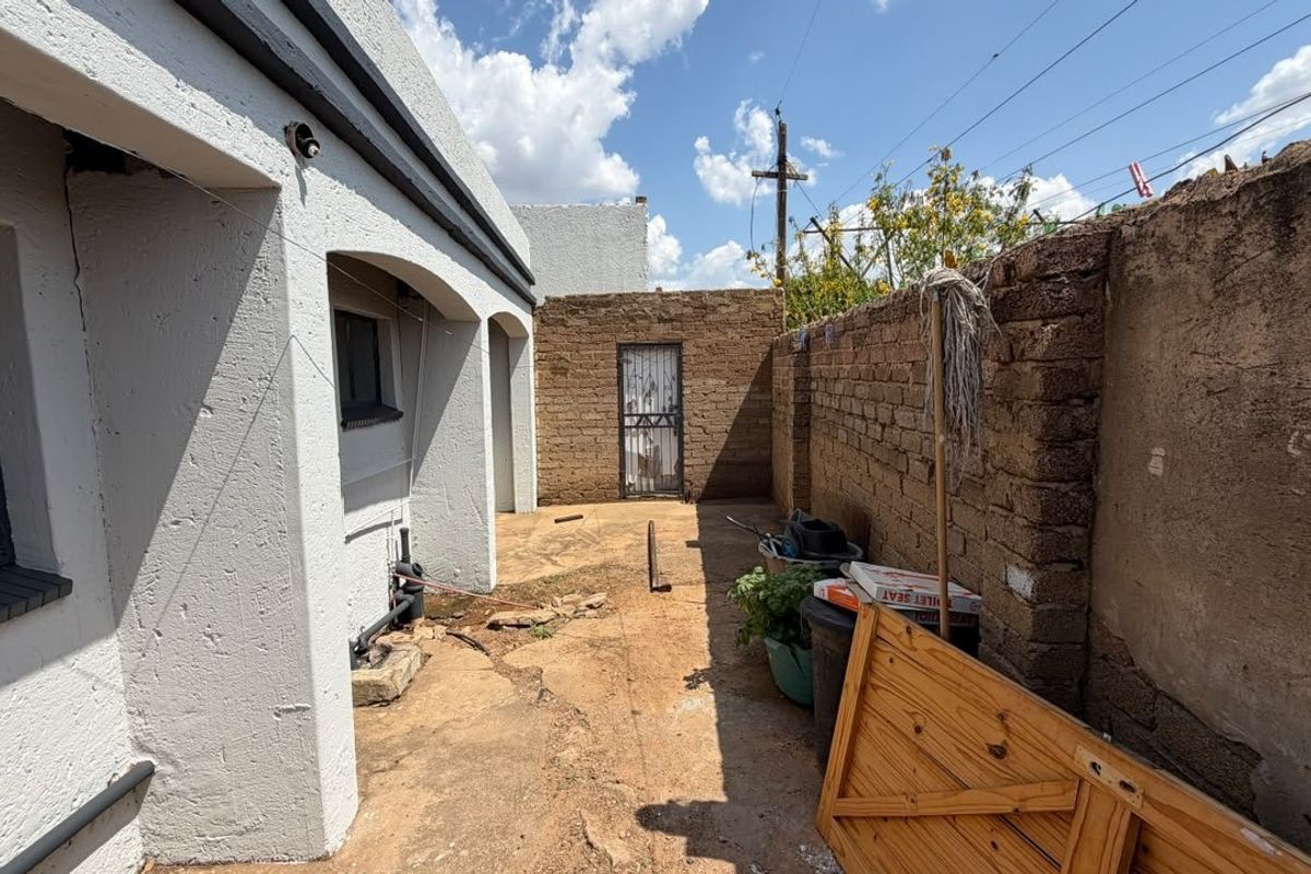 A rear exterior area featuring a brick boundary wall, concrete paving, and a security gate, requiring maintenance and landscaping improvements.